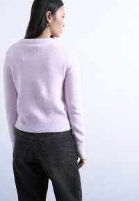 Γυναίκα με μακριά σκούρα μαλλιά που φορά χρυσούς κρίκους σκουλαρίκια, απαλό μωβ πουλόβερ και μαύρο παντελόνι ψηλής μέσης, κοιτάζοντας προφίλ μπροστά σε λευκό φόντο.