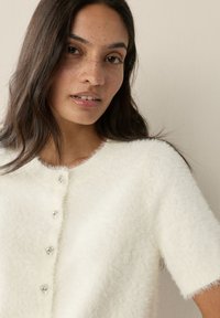 Cardigan bianco peloso con maniche corte, scollo rotondo e cinque bottoni trasparenti simili a gioielli lungo la parte anteriore per la chiusura.