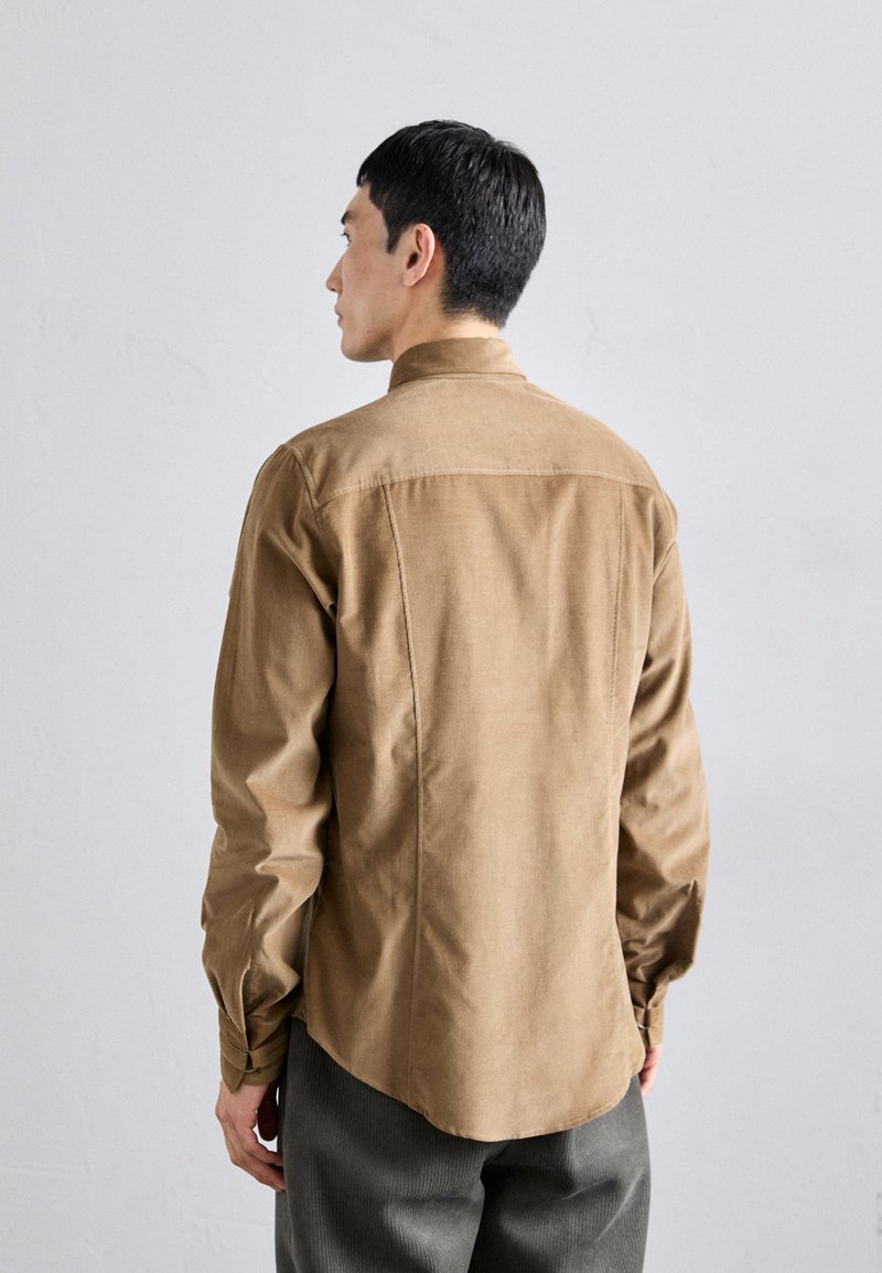 Chemise en velours côtelé marron avec un col à boutons, manches longues et ourlet arrondi. Portée sur un pantalon gris texturé.
