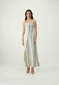 Αμάνικο, μακρύ φόρεμα σε ανοιχτό πράσινο σατέν με πλαϊνό άνοιγμα και διαγώνιες ραφές. Το μοντέλο φοράει μπεζ τακούνια και ελάχιστα αξεσουάρ.