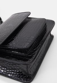 Černá strukturovaná kabelka vyrobená z lesklého materiálu s krokodýlím vzorem. Má klopový uzávěr a vrstvené kapsy s horizontálním prošíváním.