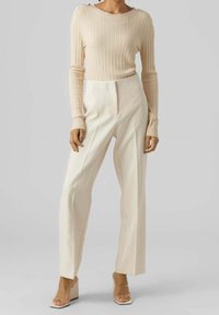 Femme portant un pull beige à manches longues côtelé, un pantalon crème ajusté taille haute et des sandales à talons blocks avec brides transparentes, se tenant sur un fond gris.