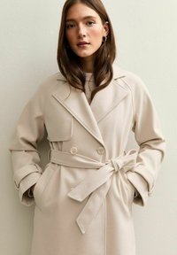 Abrigo beige de mezcla de lana con diseño de doble botonadura, solapas de pico, bolsillos delanteros y cinturón anudado en la cintura. Textura suave con una silueta estructurada.