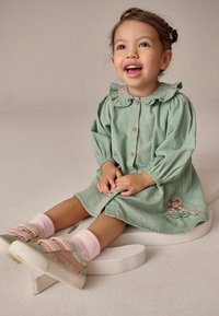 Kurzärmliges, grünes Cord-Kleid mit floralen Stickereien und Rü schekragen. Kombiniert mit Füßen in rosa Socken und pastellfarbenen Turnschuhen mit Klettverschlüssen.