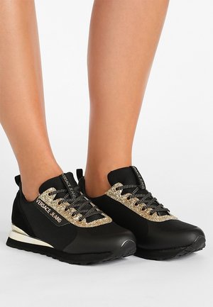 Pieds portant des baskets Versace Jeans noires avec des accents de paillettes dorées et des lacets noirs sur un fond blanc.