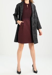 Black and white tweed coat over a burgundy dress with a textured skirt, featuring a keyhole neckline and side pockets. Black heels complete the look.