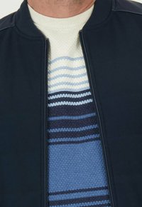 Marine bomberjack met hoge kraag, met een ritssluiting. Daaronder zit een gestructureerde trui met horizontale blauwe en crèmekleurige strepen.