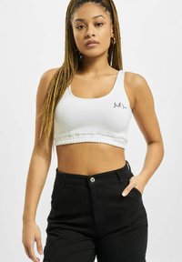 White cropped tank top with ribbed texture, featuring a stitched black logo on the chest and a gathered hem. Worn with black pants.
