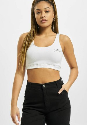 Hvit, kort tanktopp med ribbet tekstur, med en sydd svart logo på brystet og en samlet kant. Båret med svarte bukser.