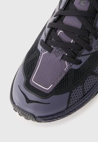 Prim-plan al unei ghete de alergare negre și violet, cu material din plasă, șireturi groase și textul "SPEEDGOAT" pe limbă.