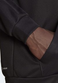 Svart atletisk hoodie med texturerad tyg, med en känguruficka och ribbade manchetter. Logotypen är placerad på den nedre vänstra ärmen.