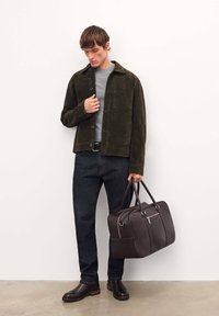 Giacca in suede verde scuro con colletto, camicia grigia, jeans scuri e borsa in pelle marrone con doppio manico e chiusura con zip. Texture liscia.