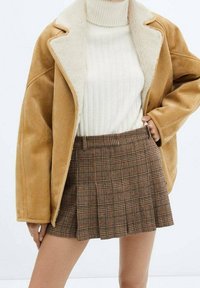 Bruine shearling gevoerde jas over een witte geribbelde coltrui. Bruine geruite geplooide minirok met een hoge taille en een getailleerd ontwerp.
