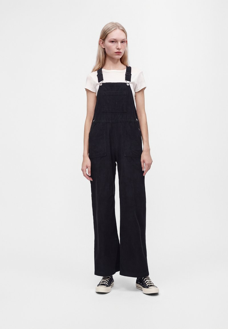 Jeune femme aux longs cheveux blonds portant une chemise blanche, une salopette noire à jambes larges et des baskets noires, debout devant un fond uni.