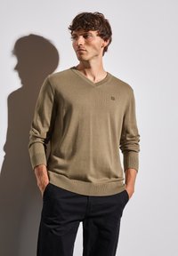 Kaki V-ringad tröja i mjukt stickat material med ribbade ärmslut och nederkant, prydd med en liten logotyp på bröstet, i kombination med mörka byxor.