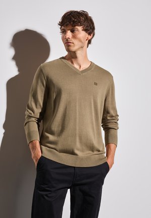 Pull en V couleur kaki en tissu tricoté doux avec des poignets et un ourlet côtelés, affichant un petit logo sur la poitrine, associé à un pantalon sombre.