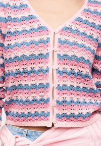 Πλεκτή ροζ, μπλε και ανοιχτό ροζ ριγέ ζακέτα με κουμπιά, φορεμένη πάνω από ανοιχτό μπλε τζιν και δεμένη με ροζ ριγέ ύφασμα.