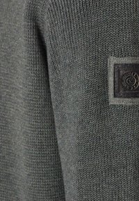Grauer gestrickter Pullover mit strukturierten Oberflächen, ausgestattet mit einem rechteckigen schwarzen Patch mit erhabenen Details auf der Brust. Weicher, mittelschwerer Stoff.