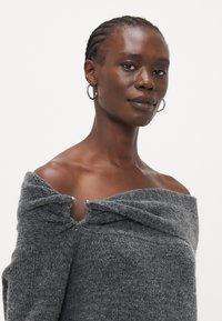 Grå off-shoulder strikkegenser med teksturert overflate, vridning ved halsen og sølvknappdetaljer.