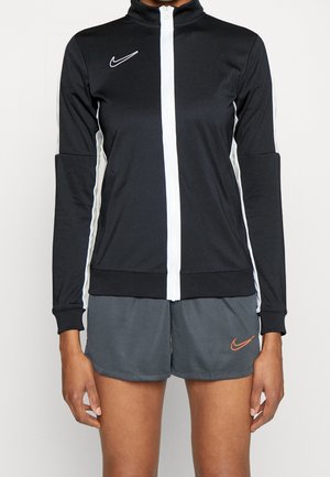 Person iført en sort og hvid lynlåstrøje og mørkegrå shorts med et lille orange Nike-logo på venstre ben.