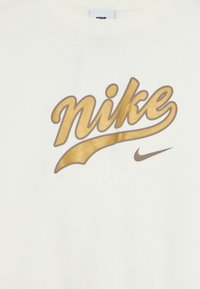 Vit skjorta med en guld- och brunt Nike-skriptlogotyp och en liten brun Nike-swoosh under bröstområdet.