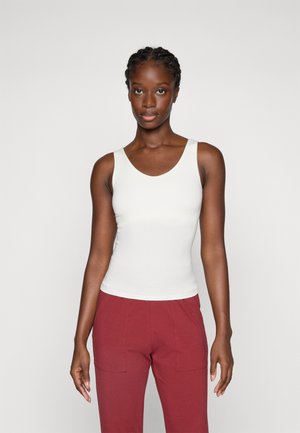 Femme avec des cheveux tressés portant un top sans manches blanc et un pantalon rouge, se tenant devant un fond gris clair uni.