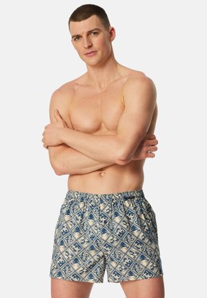Uomo con le braccia incrociate che indossa pantaloncini da bagno con motivo blu e beige, in piedi davanti a uno sfondo bianco semplice.