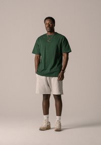 Grønn bomull t-skjorte kombinert med lysegrå shorts. Modellen har på seg brune sneakers og hvite sokker, med subtile merkevareelementer.