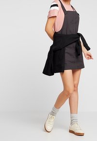 Grå denimklänning med axelband, lager ovanpå en rosa randig skjorta. Kombinerad med vita sneakers och grå strumpor, en svart tröja knuten runt midjan.