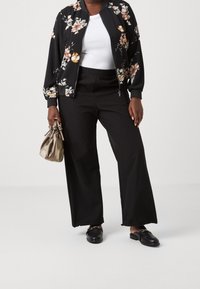 Černá květinová bomber bunda přes bílý top, doplněná o široké černé kalhoty a černé slip-on boty. Drží malou, staženou bronzovou kabelku.