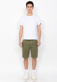 Camiseta de algodón blanca de manga corta, pantalones cortos verde oliva y zapatillas beige. Cuenta con una etiqueta negra en la manga de la camiseta.
