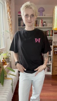Person med kort blondt hår iført en sort t-shirt med et pink sommerfuglelogo, hvide bukser og tatoveringer på venstre arm i et stue.
