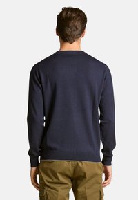 Jersey de punto azul marino con mangas largas y cuello redondo, con puños y dobladillo acanalados, llevado sobre pantalones caqui.