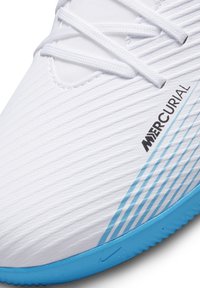 Valkoinen jalkapallokenkä teksturoidulla päälliosalla, vaaleansinisillä yksityiskohdilla ja Nike-logoilla. Ominaisuuksiin kuuluvat vaakasuorat raidat sivuilla ja nauhoissa.