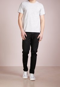 T-shirt grigia in cotone a maniche corte abbinata a jeans neri slim-fit e sneakers bianche, esposta su uno sfondo rosa chiaro.