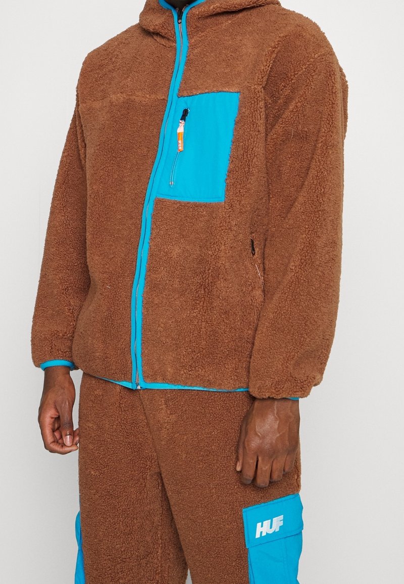 Veste en polaire marron et pantalon avec des accents bleu vif et des poches zippées, portés par une personne debout à la peau foncée.