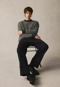 Grauer, zigzagförmiger Strickpullover, schwarze weit geschnittene Hose und schwarze Schuhe. Modell sitzt auf einem schwarzen Stuhl mit verschränkten Armen.