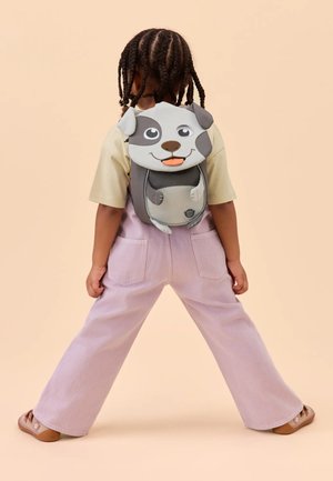 Child with braided hair wearing a dog-shaped backpack, beige shirt, light purple pants, and sandals, standing with legs apart on beige background.