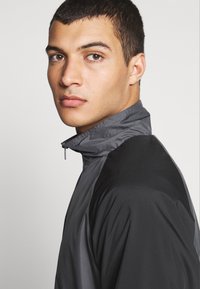Water-resistant, gray jacket with a stand-up collar and contrasting black accents. Features a smooth texture and zip closure.