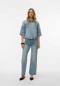 Džinsa jaka un džinsi komplekts, gaiši zils ar kniedēm, īss jaka modelis, pārlieku platām piedurknēm, taisnas kājas bikses ar nelielu nodilumu un melni apavi.