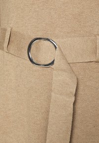 Beige stickad tyg med ett brett bälte som har en silverfärgad, cirkulär spänne. Bältet har en texturerad yta och är fäst vid plagget.