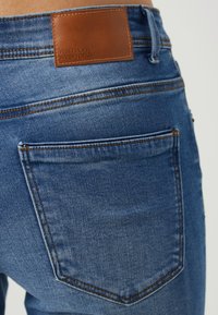 Džínové kalhoty střední modré barvy s texturovaným povrchem, s koženou etiketou na pase a kontrastním prošitím na kapsách.