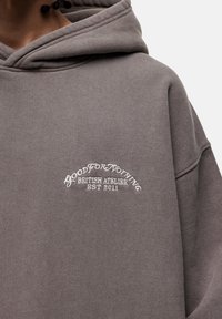 Felpa grigia chiara con grande tasca a marsupio, cordino regolabile e testo ricamato che legge "GOOD FOR NOTHING BRITISH ATELIER EST 2011."