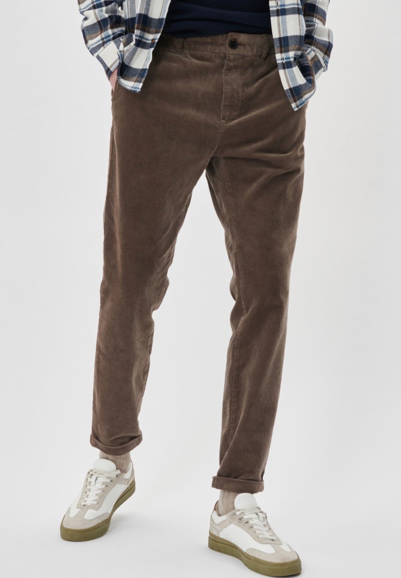 Pantalones de pana marrones con corte recto y ligero estrechamiento, con cierre de botón y puños doblados, combinados con zapatillas blancas.