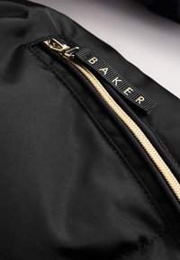 Primer plano de una tela negra con una cremallera dorada y una lengüeta negra con la etiqueta "BAKER" en letras doradas.