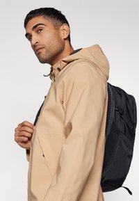 Beige wasserdichte Jacke mit verstellbarem Kapuze; verfügt über eine Reißverschlusstasche und einen schwarzen Rucksack mit strukturierten Akzenten. Modell posiert seitlich.
