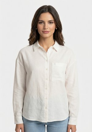 Giovane donna con lunghi capelli scuri che indossa orecchini di perle, una camicia di lino bianca con bottoni e taschino sul petto, e jeans azzurri, in piedi contro uno sfondo bianco.