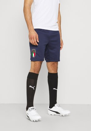 Homme portant un short de football bleu marine Italie, des chaussettes noires Puma montantes et des crampons blancs Puma, debout devant un fond clair uni.
