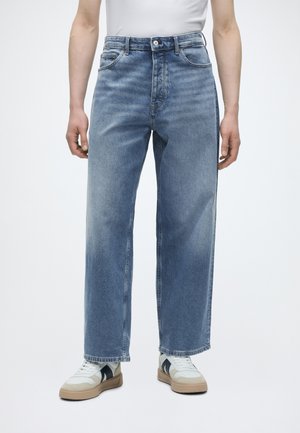 Άτομο με ανοιχτό μπλε τζιν ευθείας γραμμής και λευκά αθλητικά παπούτσια με μαύρες ρίγες, στέκεται μπροστά σε απλό λευκό φόντο.