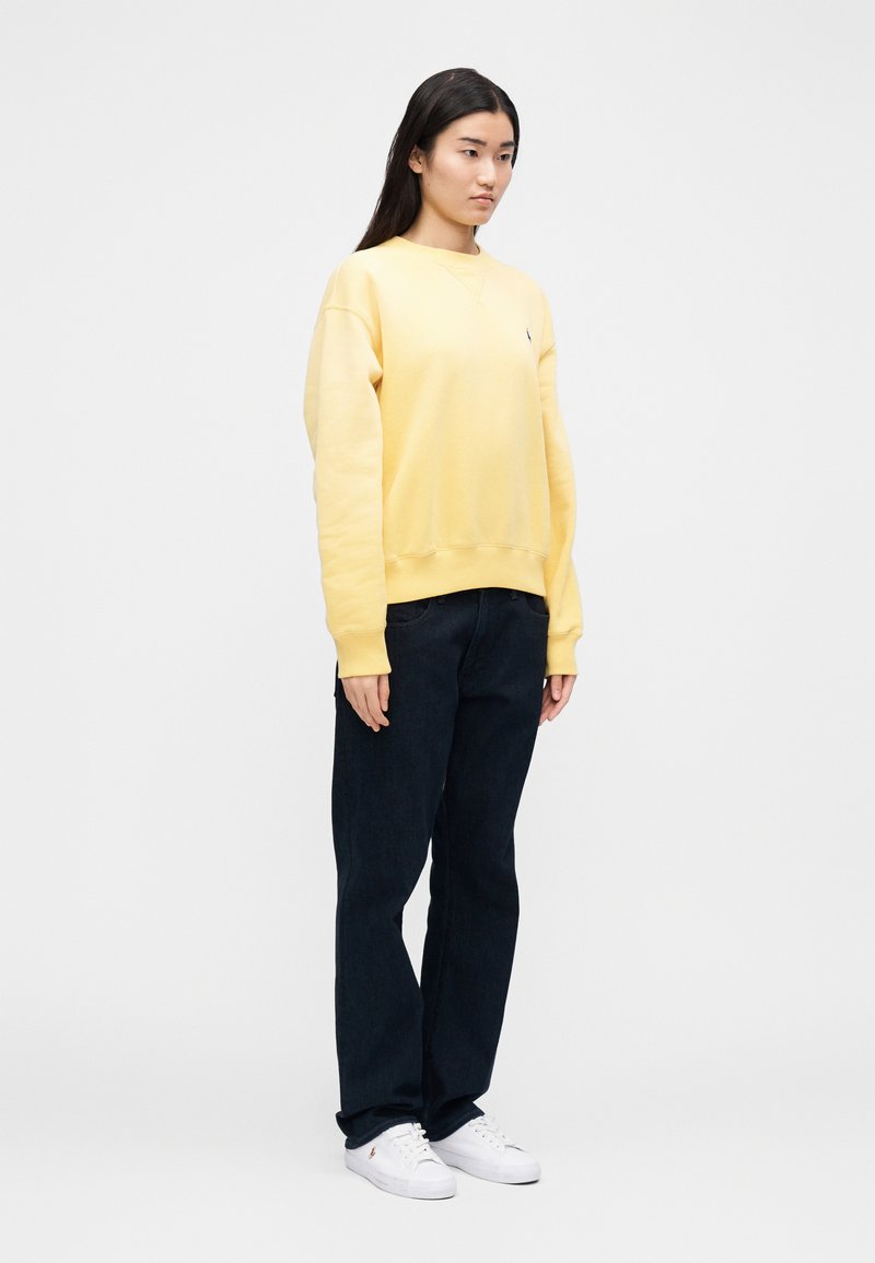 Sweat-shirt jaune avec un col rond et des poignets côtelés, associé à un jean bleu foncé et des baskets blanches. Design simple, aucun motif visible.